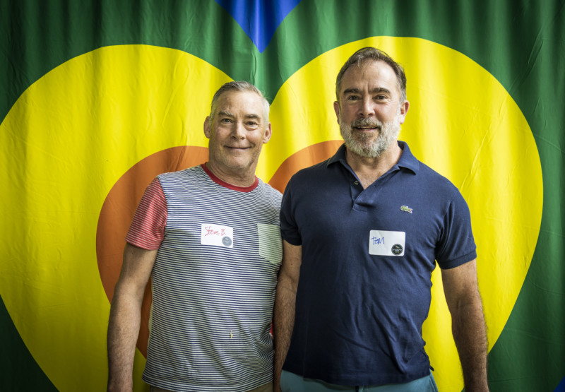 Two men in front of a rainbow-colored heart.