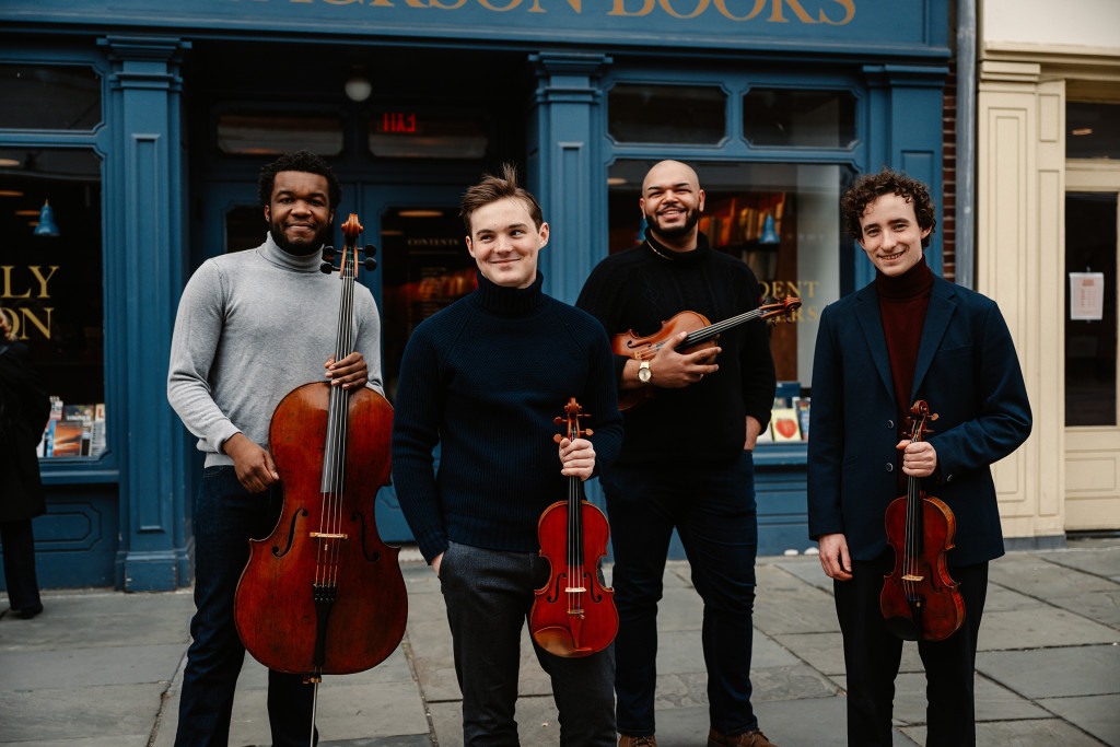 Members of the Isidore String Quartet pose with their instruments on a city sidewalk in front of a storefront.