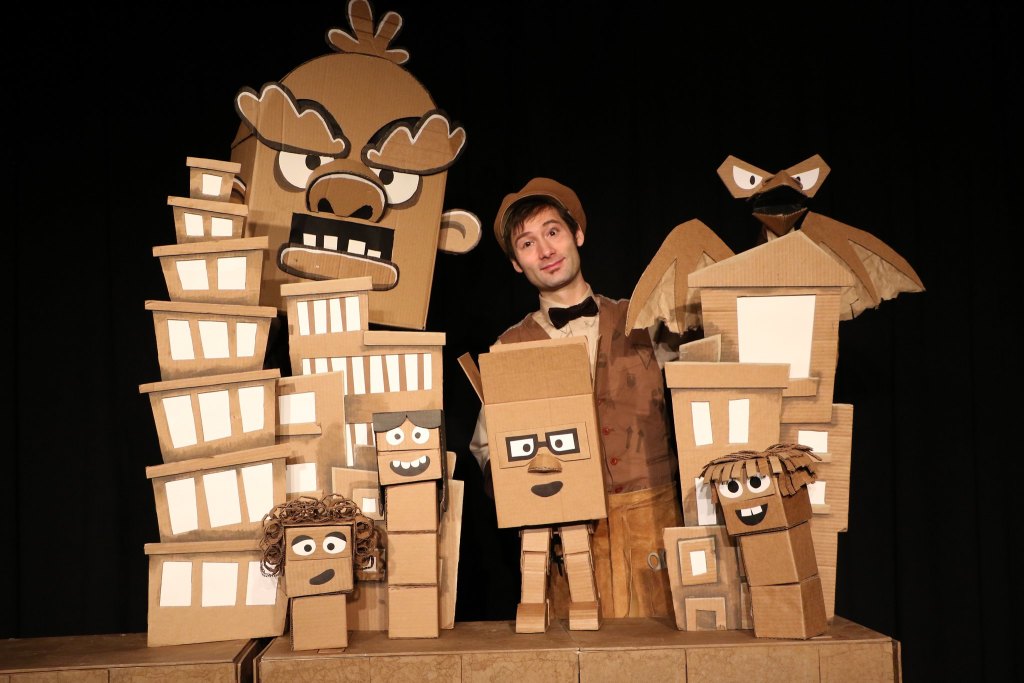 Puppeteer Brad Shur, dressed in a brown suit with a bowtie and a brown hat to match the color of his cardboard puppets, poses with several of his puppets, which have unique looking faces and are about as tall as he is.