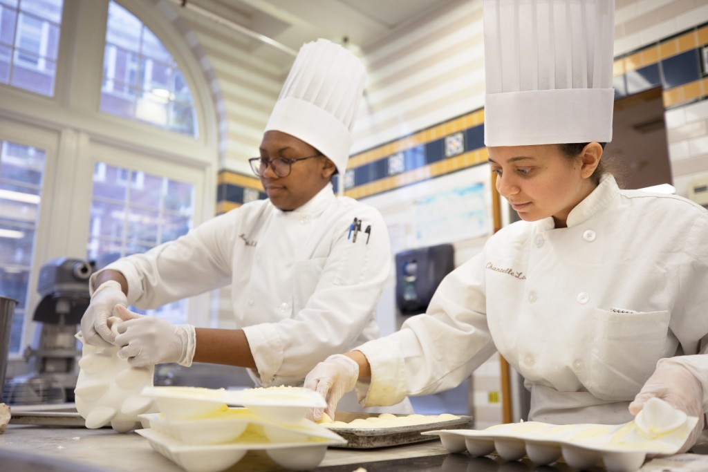 Two chefs, wearing all-white chef's attire and tall chef's hats, prepare a meal in the kitchen at the Culinary Institute of America in Hyde Park.