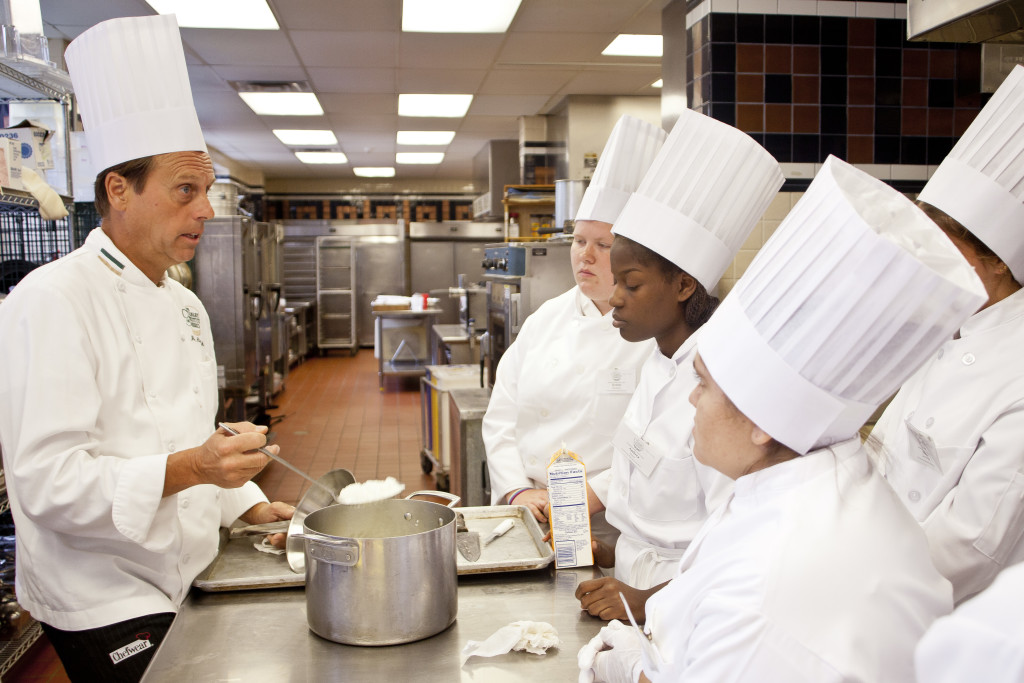 Student chefs gather around an instructor demonstrating a cooking technique in the kitchen at the Culinary Institute of America in Hyde Park