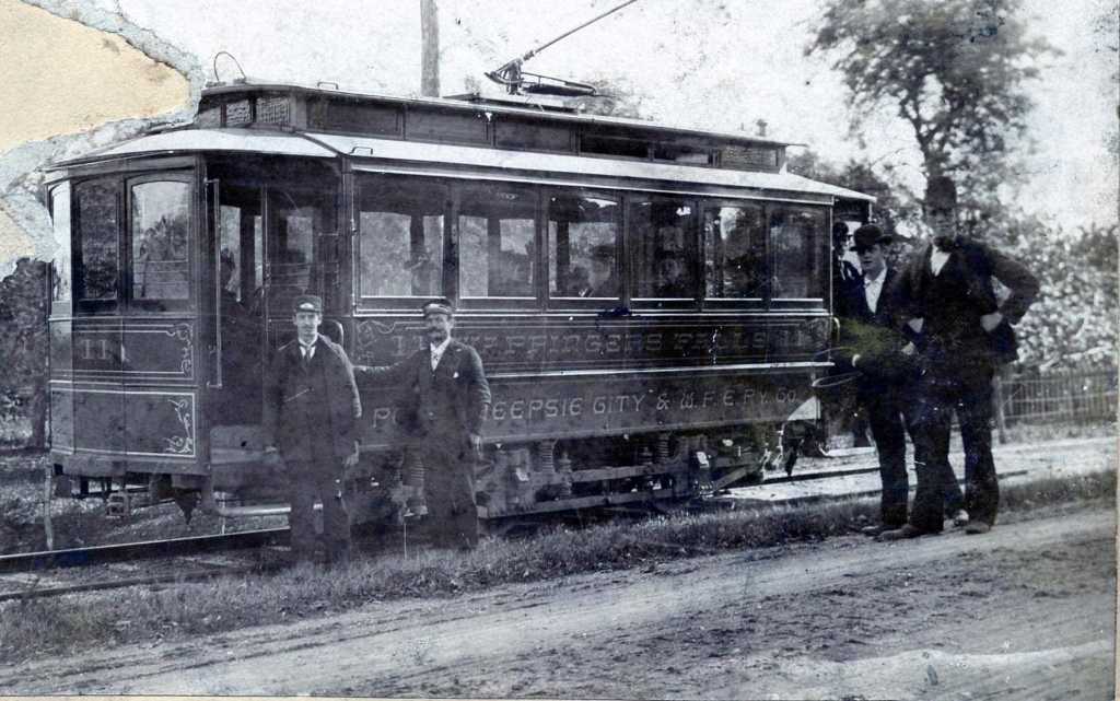 Black and white, grainy photo of an old trolly car with two conductors standing and two pedestrians walking in front of it.