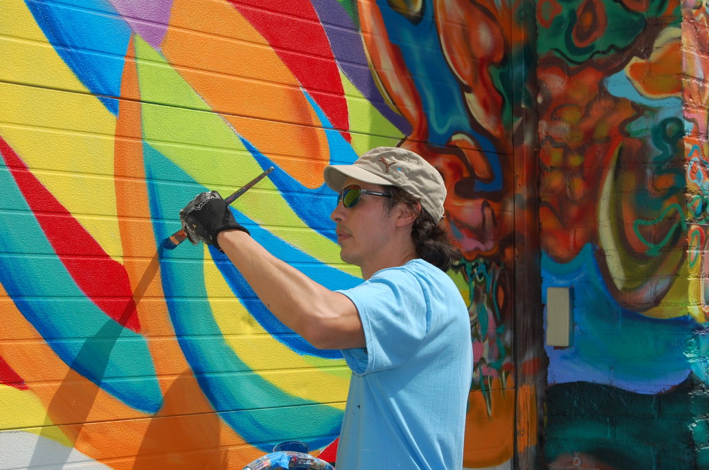 A young male painter with shoulder length dark hair, a beige baseball cap, sunglasses and a light blue tee shirt paints a colorful mural on a brick wall in Poughkeepsie.