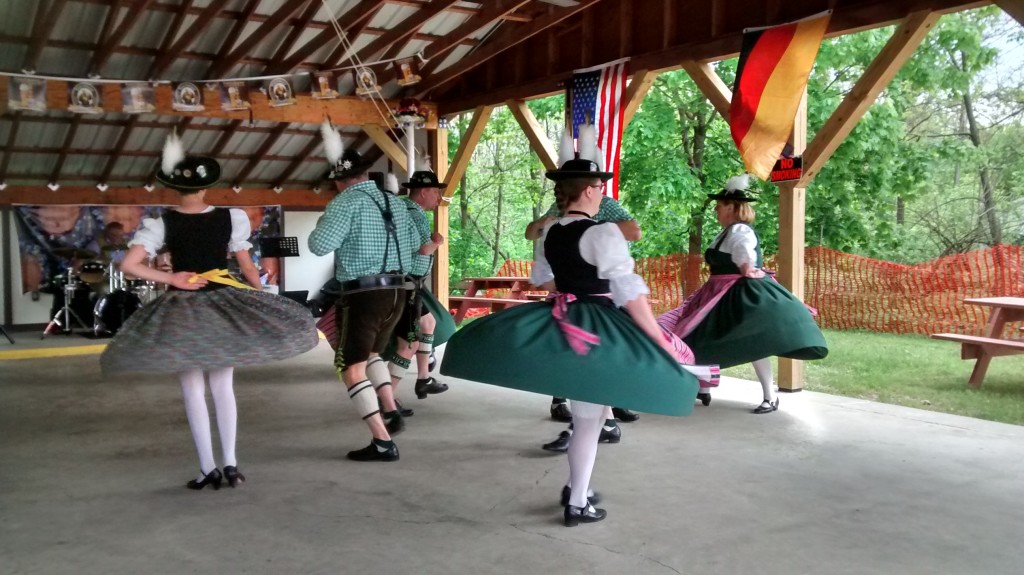 A group of male and female dancers, the males wearing black and green lederhosen and the women wearing black and green swirling dresses, dances under a pavilion during an Oktoberfest celebration at Germania of Poughkeepsie.