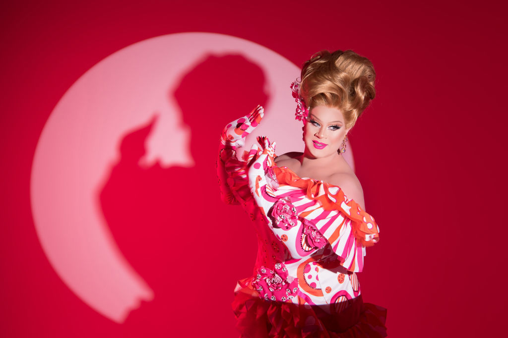 With her big, poofy hair and bright pink and orange outfit, drag queen Nina West poses in the spotlight with a bright pink background behind her.