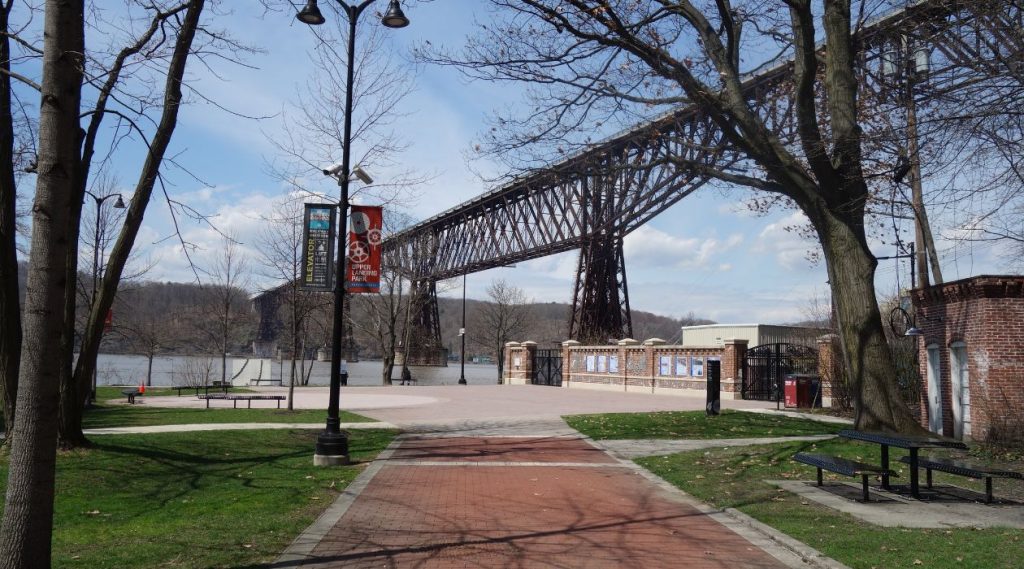 Upper Landing Park on the Hudson River in Poughkeepsie, with its green grass, brick walkways, ornate lampposts and the Walkway Over the Hudson towering in the background against a blue sky.