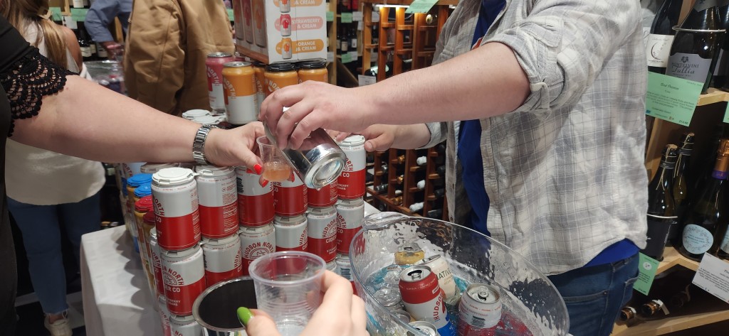 Outstretched hands holding empty, clear-plastic cups reach toward another outstretched hand pouring a can of cider from behind a display table holding stacks of red and white cans at Boutique Wines, Spirits and Ciders in Fishkill.