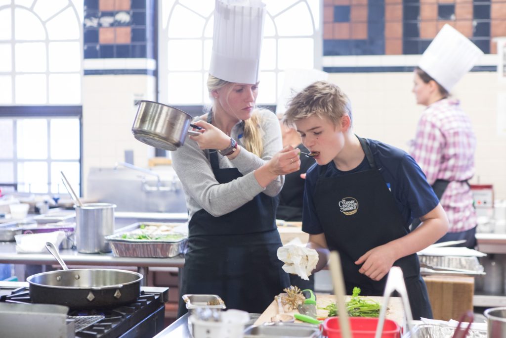 A female instructor with blonde hair tucked up under her tall, white chef hat, a gray shirt and a black apron holds a spoon out for an adolescent male student with shaggy blonde hair and a black shirt and apron to taste during a cooking class at the Culinary Institute of America.