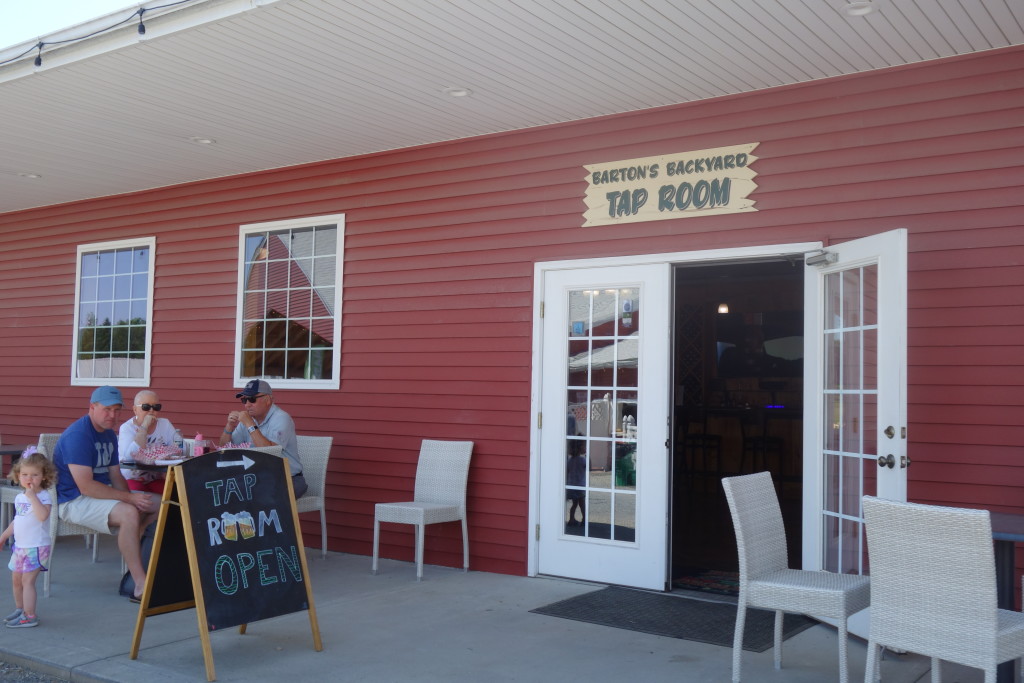 The white double doors, the right one of which is propped open, at the front of the Barton Orchards taproom, a red building with tables and chairs outside and an A frame chalkboard.