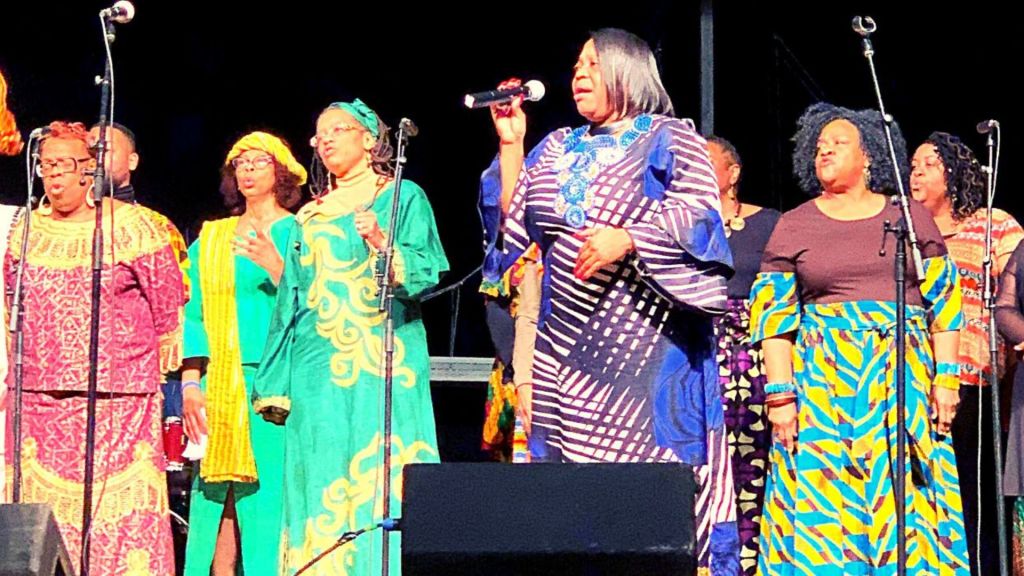 A line of six female gospel singers wearing colorful, flowing gowns performs on stage at the Hudson Valley Gospel Festival.