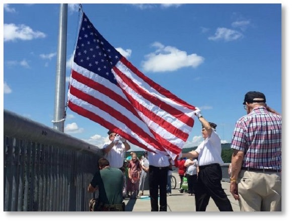 Three men in dress military uniforms change the flag on the flagpole at the center of the Walkway Over the Hudson as several other men salute.