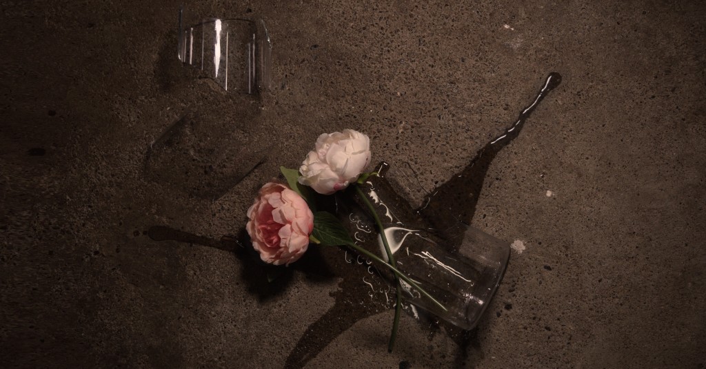 A clear glass vase containing two flowers has fallen on a dark gray cement floor and broken.