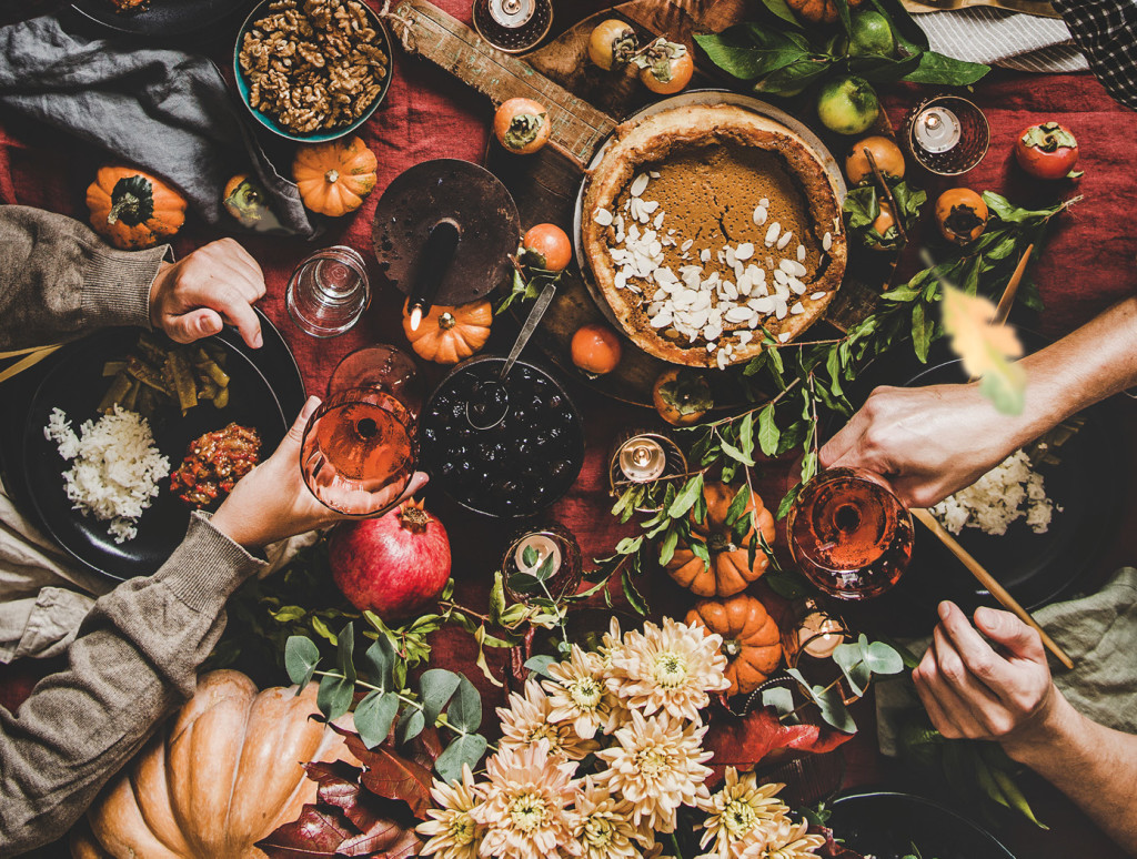 An overhead view of a festively decorated Thanksgiving table with classic favorites, including pumpkin pie, as hands clutching wine glasses reach out to toast the holiday.