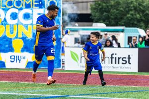 A youth and adult player for Westchester Soccer Club run out onto the field together.