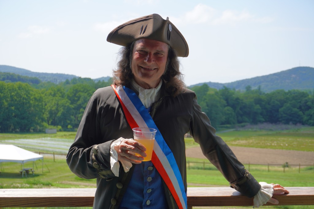 Storyteller Jonathan Kruk, dressed in Colonial-era garb and holding a cup of Treasury Cider, stands on the covered porch at Fishkill Farms in Hopewell Junction during the Revolutionary Cider Celebration.