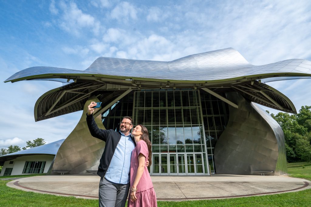 a man and woman take a selfie in front of a large, metallic building with a wavy roof resembling a bird or a wave. It is the Fisher Center at Bard College designed by Frank Gehry