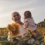 a grandfather in a white shirt smiles with joy while holding his small granddaughter surrounded by bright yellow sunflowers at Barton Orchards