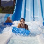 three visitors to SplashDown Beach race down a blue waterslide on blue sled mats. the woman centered has beaten her husband (left) and son (right) in the slide race