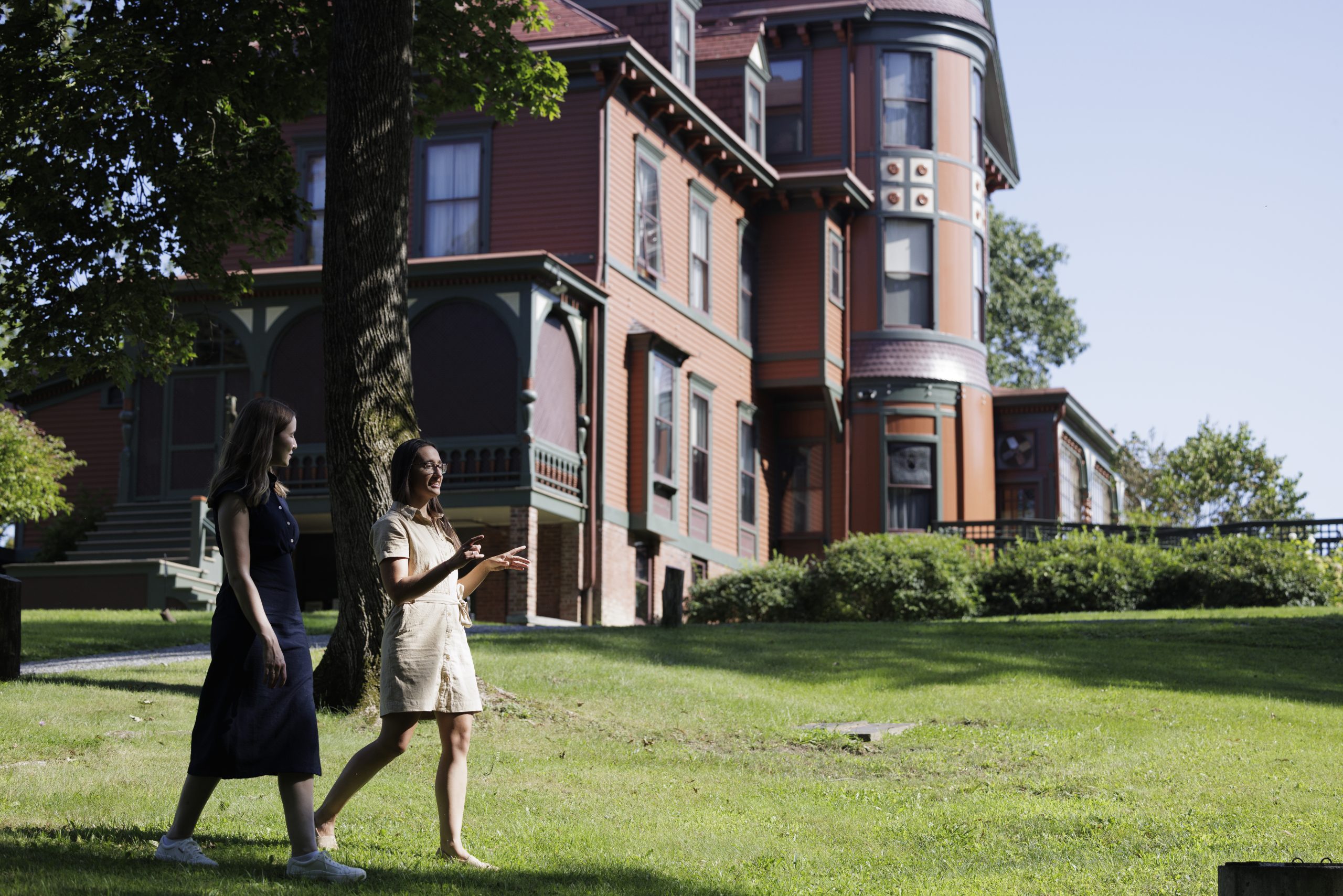 two women cross in front of a tall Victorian mansion the Wilderstein Estate which is a faded maroon color