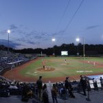 a wide shot of a minor league baseball field at dusk with stadium lights illuminating the field