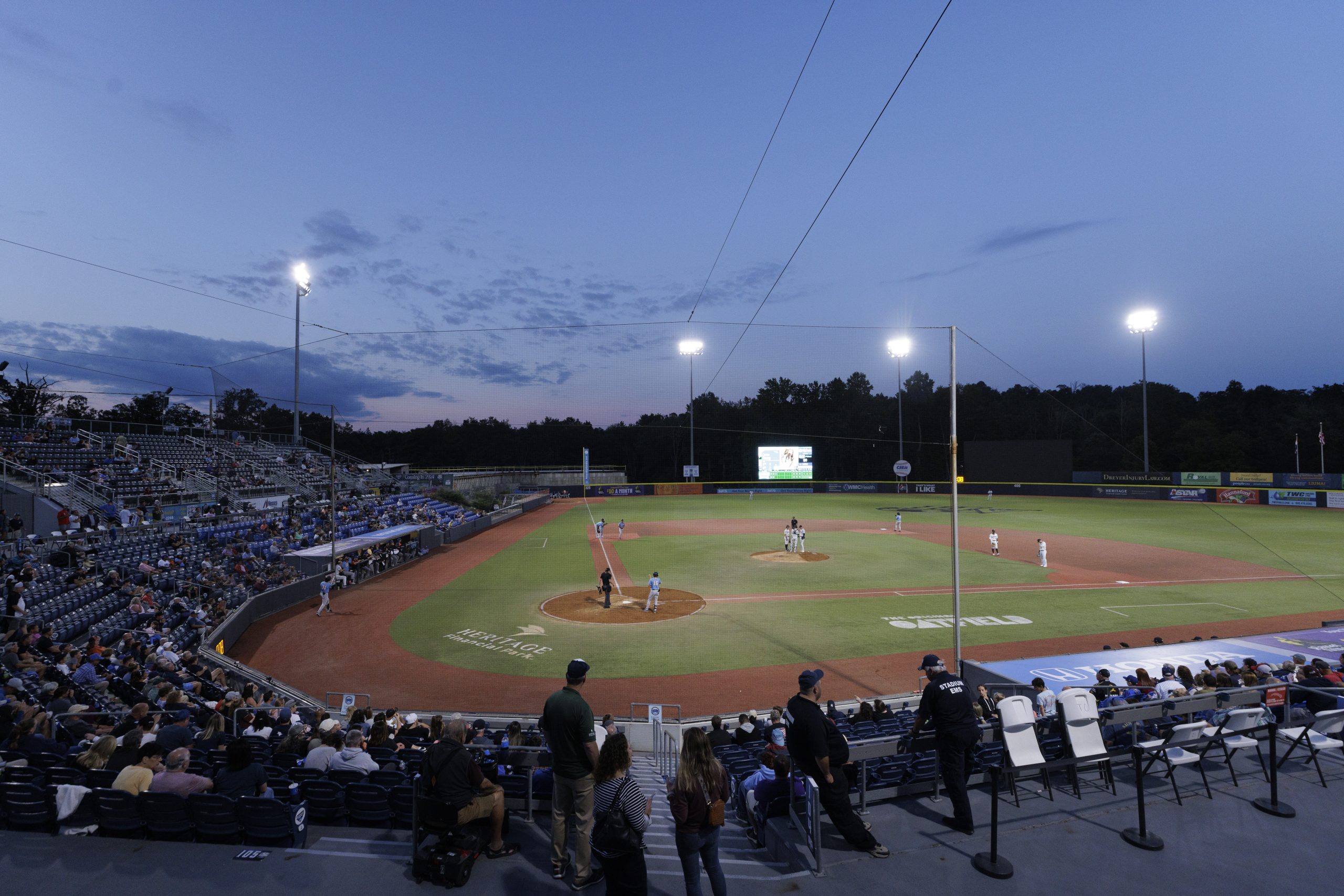a wide shot of a minor league baseball field at dusk with stadium lights illuminating the field