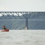 a woman in sunglasses paddles a red canoe on the blue Hudson River with a view of a bridge behind her, the Walkway Over the Hudson, marked by its many interconnected lines