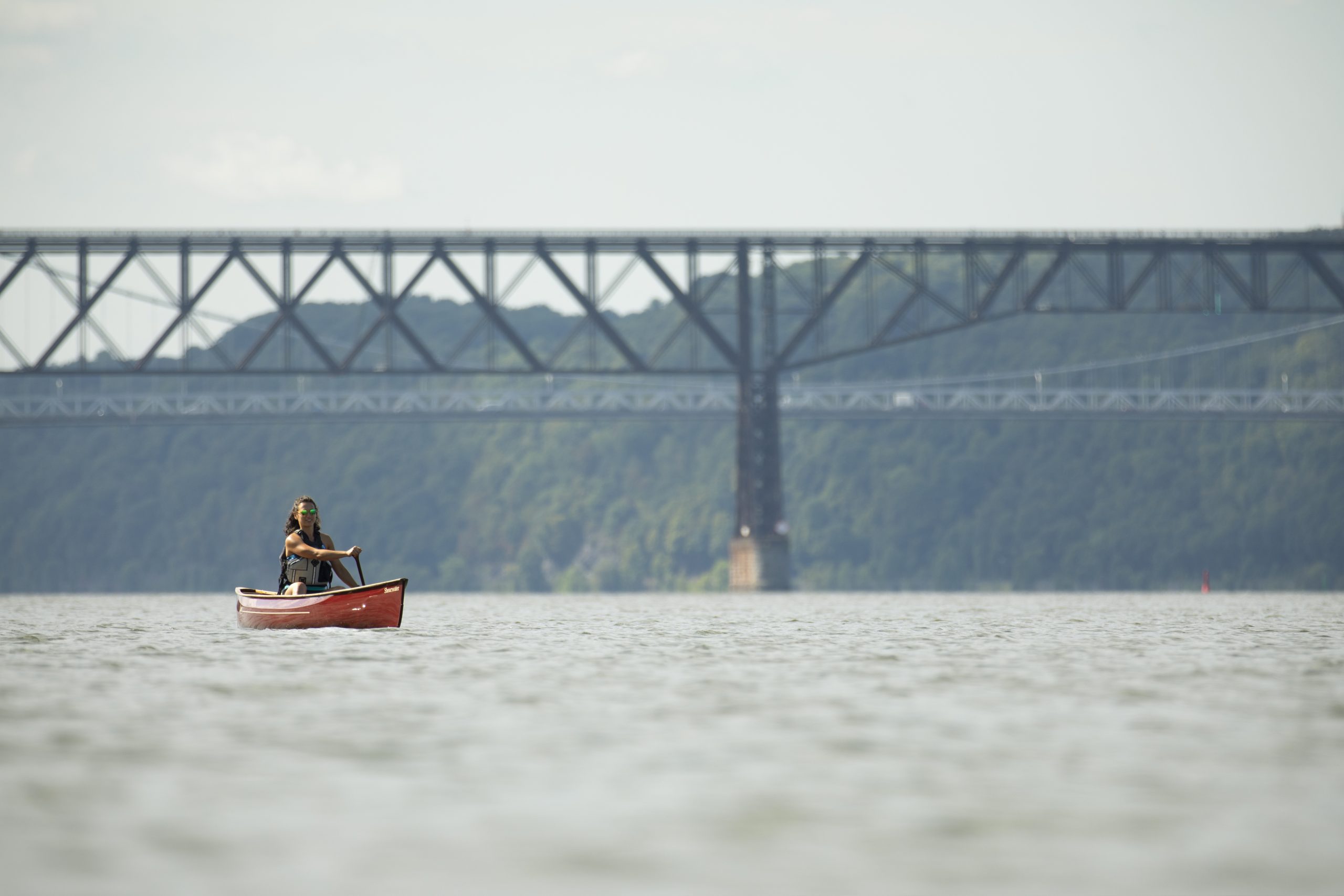 a woman in sunglasses paddles a red canoe on the blue Hudson River with a view of a bridge behind her, the Walkway Over the Hudson, marked by its many interconnected lines