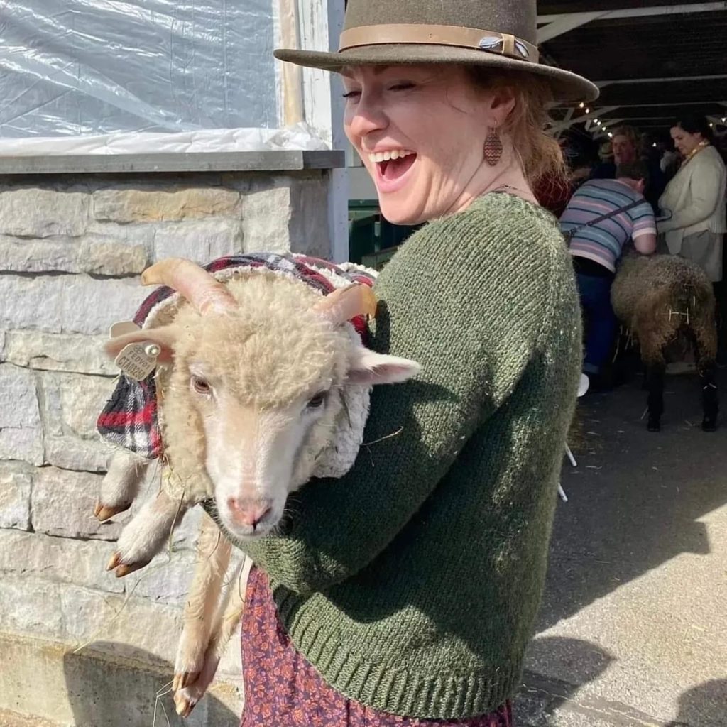 A young woman wearing a wool sweater smiles as she holds a sheep in her arms at the New York State Sheep and Wool Festival at the Dutchess County Fairgrounds in Rhinebeck